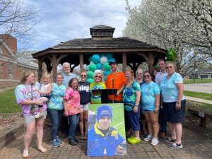 Rosie Hershberger is standing with a group of people in front of a pavilion behind her son, Christopher's photo.